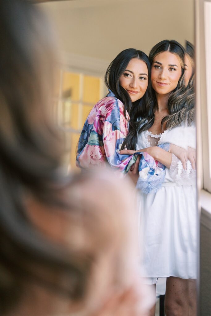 Two women stand in front of a mirror. One woman is wearing a floral robe, and the other is in a white outfit. They are gazing at their reflection together.