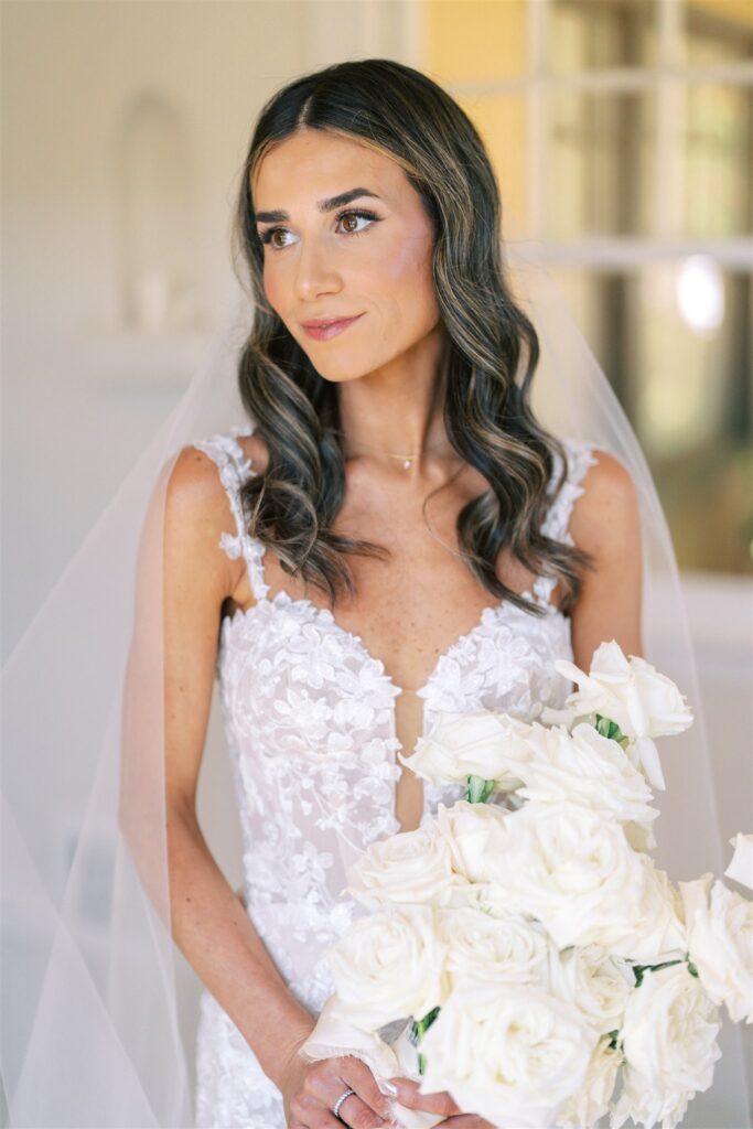 Bride in a lace wedding dress holds a bouquet of white roses, standing indoors with a soft background.