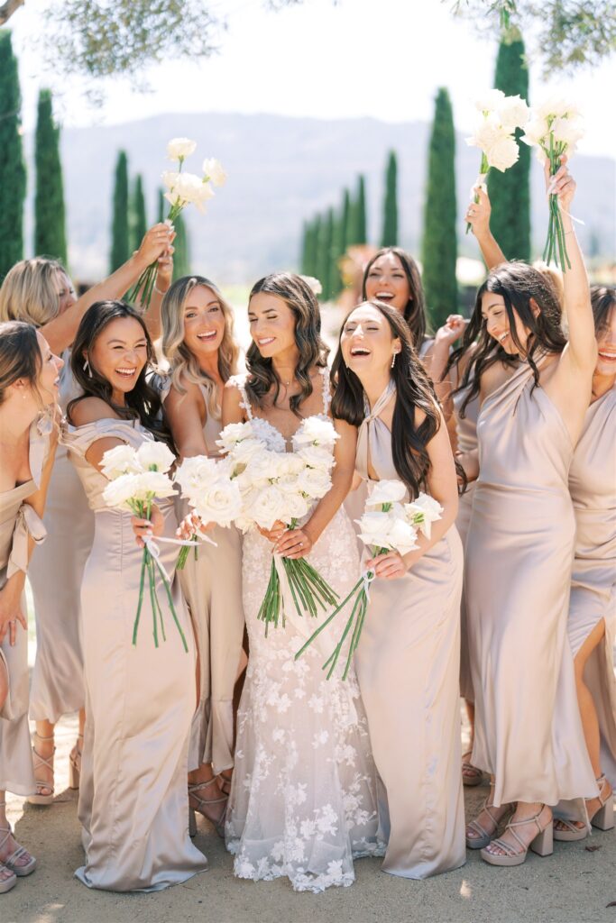 A bride surrounded by bridesmaids, all in light beige dresses, hold white bouquets and smile outdoors with tall trees in the background.