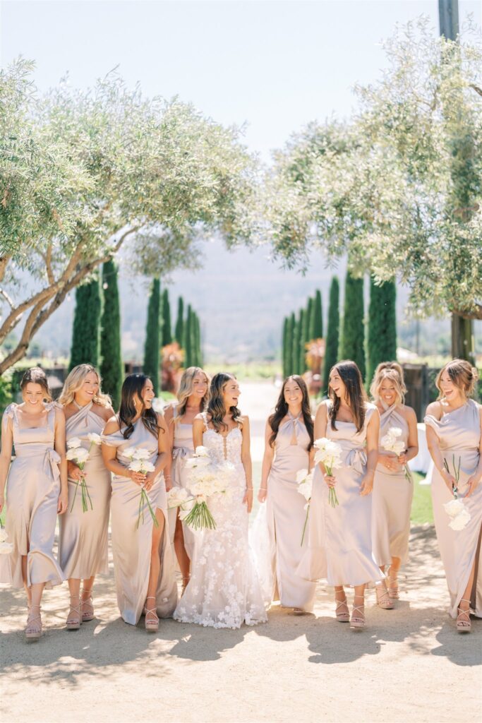 A bride in a white dress stands with seven bridesmaids in light dresses, holding white flowers, outdoors under a clear sky with trees lining the path.