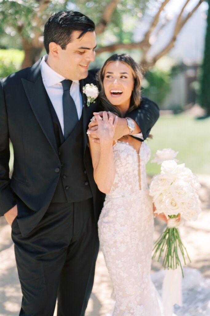A bride and groom stand outdoors, smiling. The bride holds a bouquet of white flowers while the groom has his arm around her shoulder. They are in formal wedding attire.