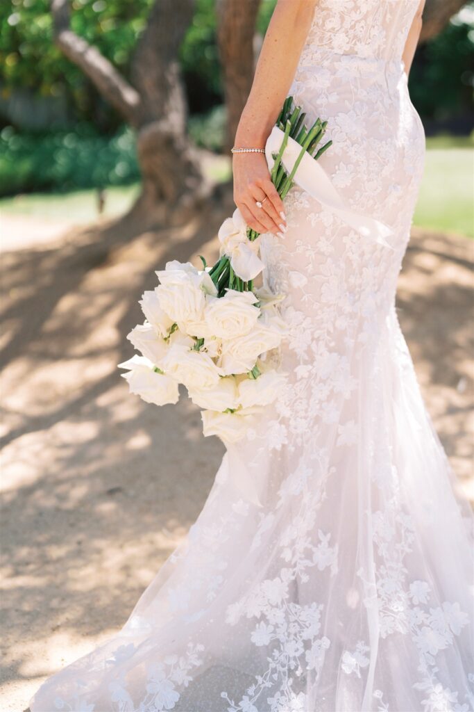 Bride in a lace gown holding a bouquet of white roses outdoors.
