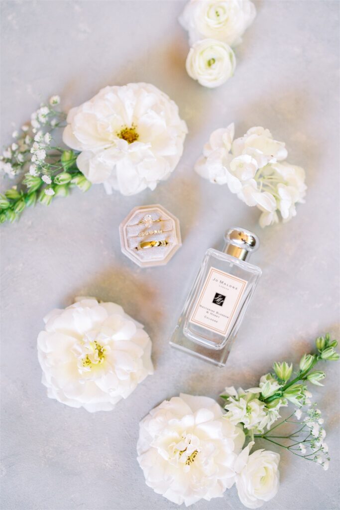Perfume bottle surrounded by white flowers and a velvet ring box with two rings, placed on a light gray surface.