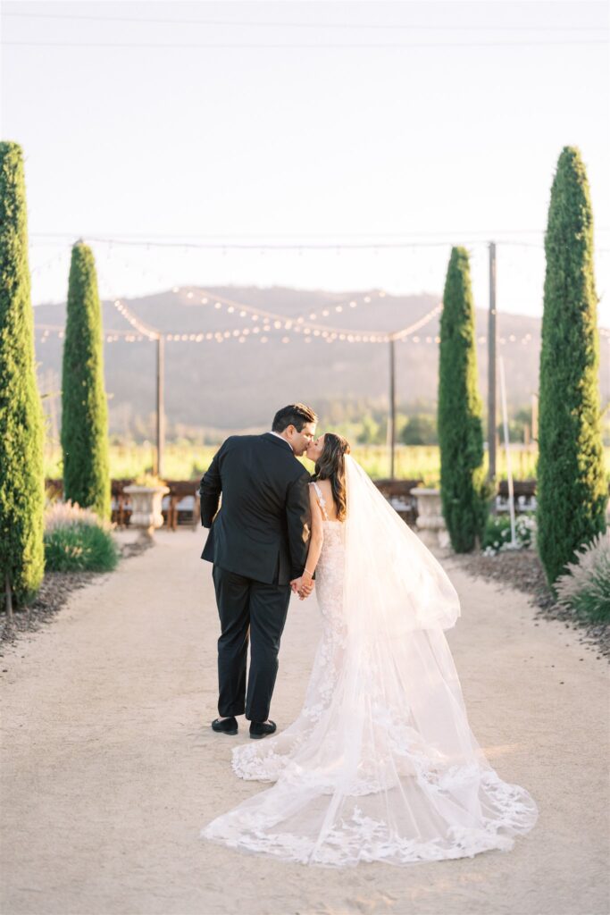 A couple in wedding attire walks along a path, holding hands, surrounded by tall trees and lights.