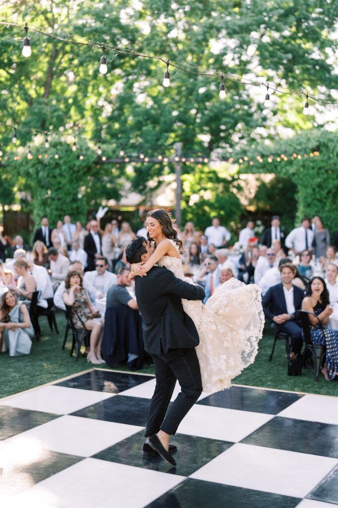A bride and groom dance on a checkered floor outdoors, surrounded by seated guests. String lights hang above, and trees provide a green backdrop.