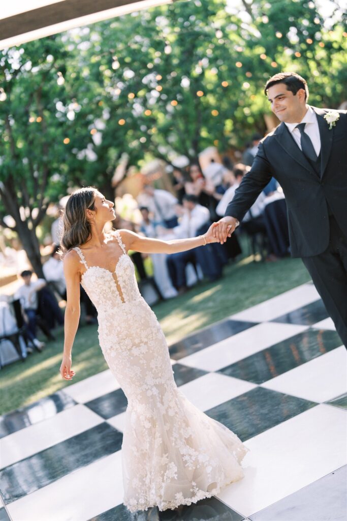 A couple dances together on a black and white checkered floor outdoors, surrounded by guests and greenery.