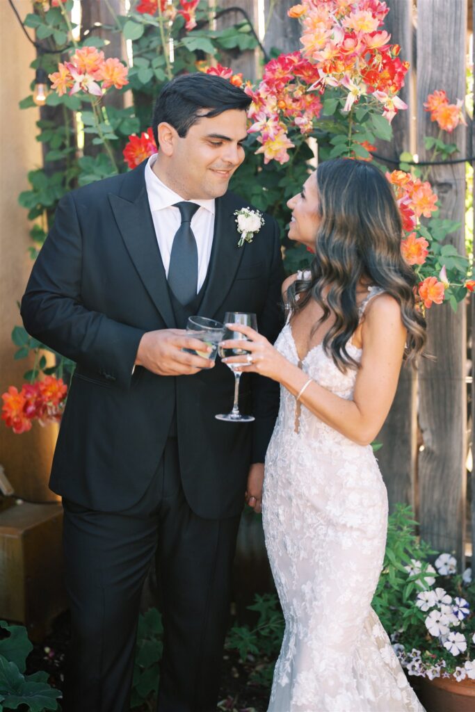 A couple in formal attire stand outdoors, holding wine glasses. The man wears a black suit, and the woman wears a white lace dress. They are surrounded by vibrant flowers.