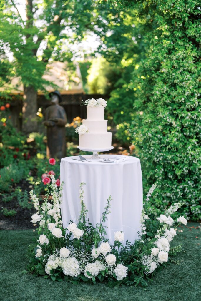 A white three-tiered cake on a pedestal table, surrounded by white and green floral arrangements in a garden setting.