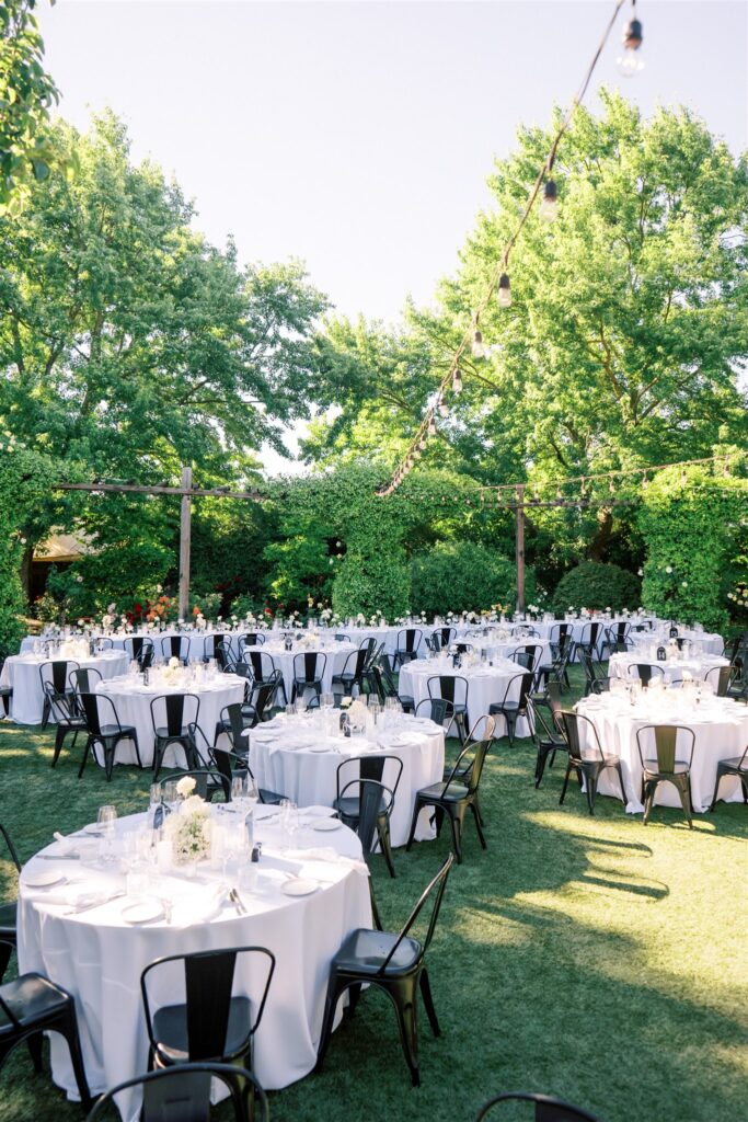 Outdoor event setup with round tables covered in white tablecloths, black chairs, and string lights above. The setting is surrounded by lush greenery.