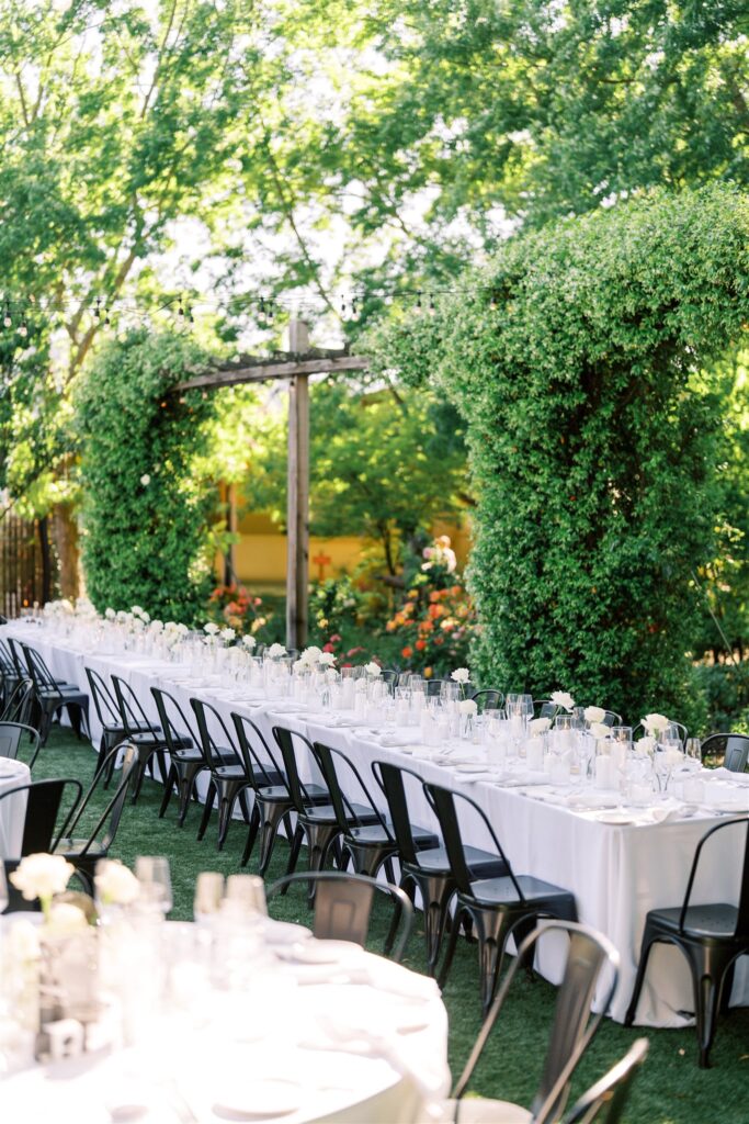 Long tables with white tablecloths and black chairs set in an outdoor garden with lush greenery and flowers.