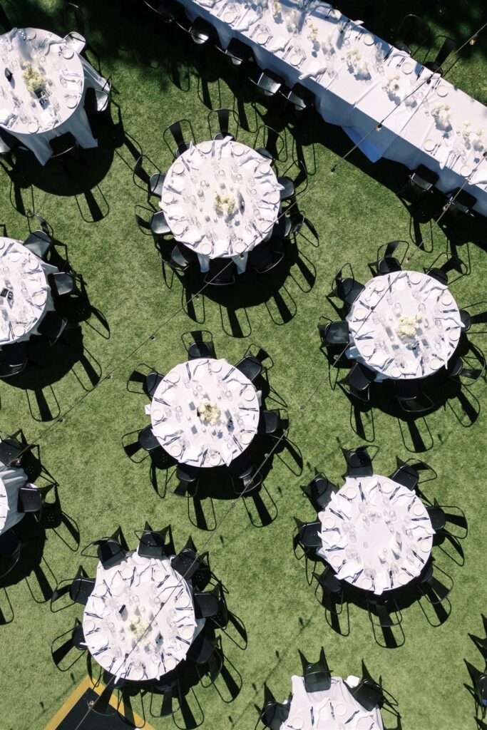 An aerial view of multiple round tables set for an outdoor event on a grassy area, with white tablecloths, black chairs, and floral centerpieces.
