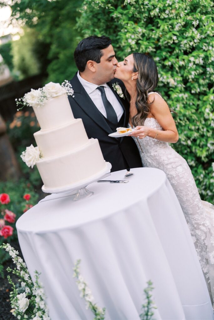 A couple kisses in front of a three-tiered white wedding cake on a table outdoors. The bride holds a plate with a slice of cake.