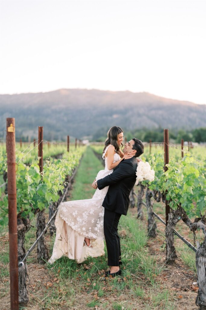 A groom lifts a bride in a vineyard, both smiling, with a mountain in the background.