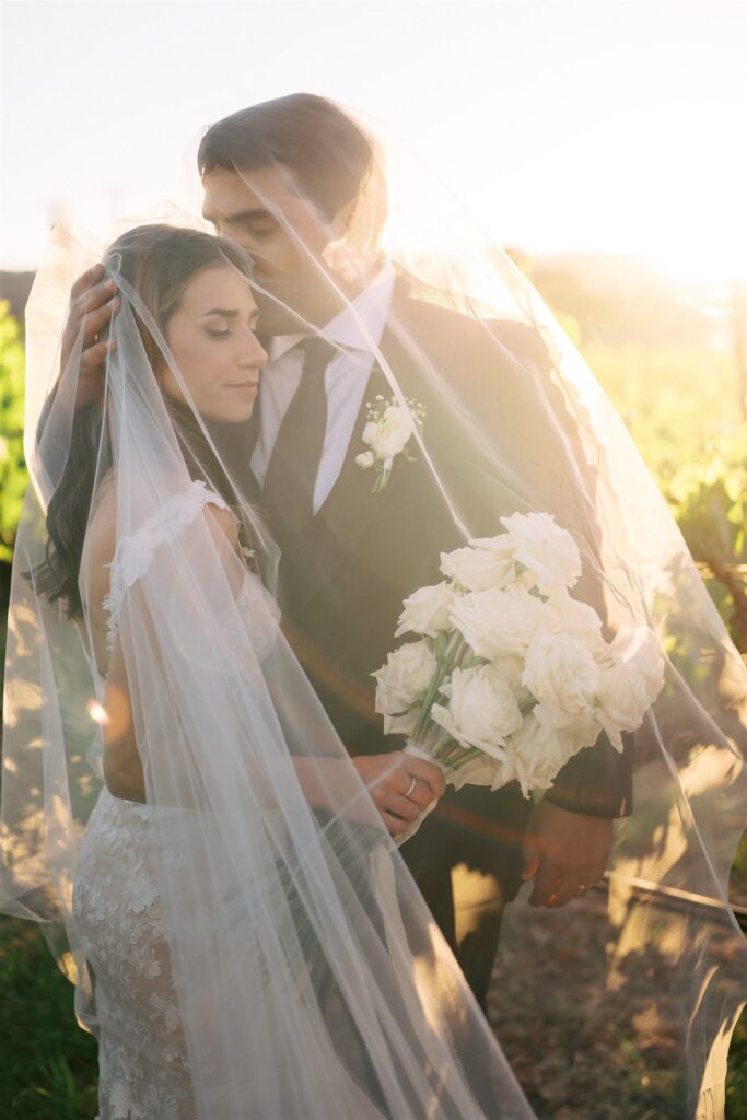 A bride and groom stand under a sheer veil in an outdoor setting. The bride holds a bouquet of white roses.