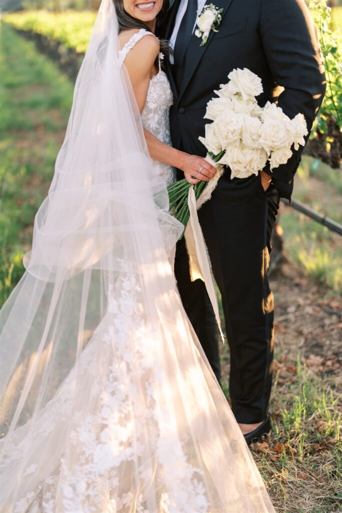 Bride and groom in a vineyard, with the bride wearing a lace gown and holding a bouquet of white roses.