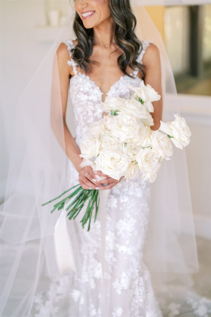 Bride in a lace wedding dress holds a bouquet of white roses, with a veil draped over her hair.