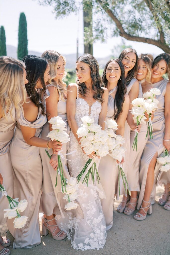 A bride in a white gown stands with bridesmaids in taupe dresses, all holding white rose bouquets, outdoors.