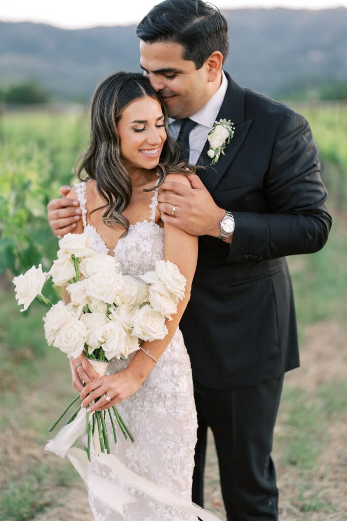 A couple in formal attire poses outdoors. The woman holds a bouquet of white roses, and the man embraces her from behind. They are in a natural setting with greenery in the background.