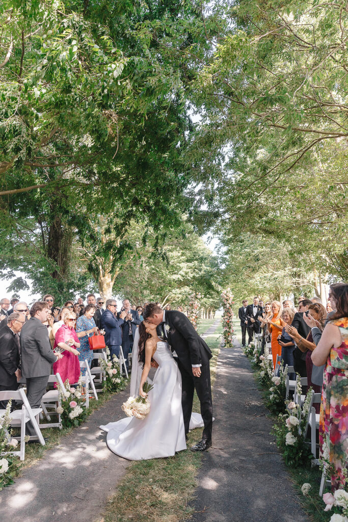 A couple kisses at the end of an outdoor wedding aisle, surrounded by guests seated in white chairs under a canopy of trees.