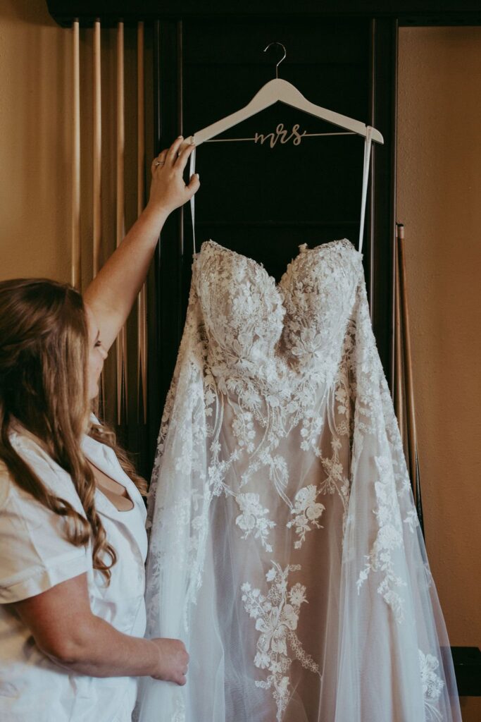 A person holds up a lace wedding dress on a hanger labeled "mrs," showcasing the detailed floral patterns on the gown.