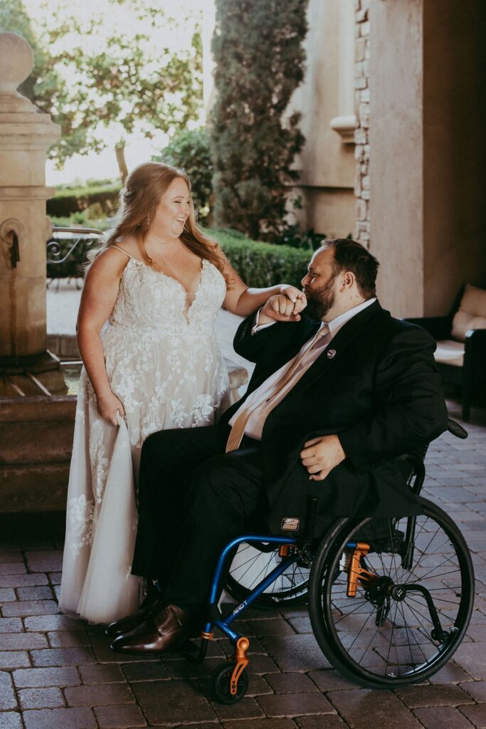 A bride in a white dress and a groom in a suit, seated in a wheelchair, share a joyful moment outdoors near a fountain.