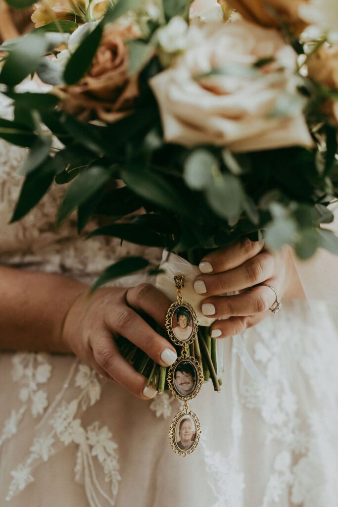Close-up of a bride holding a floral bouquet with three photo lockets hanging, each displaying a portrait. The bride wears a lace dress and has painted nails.