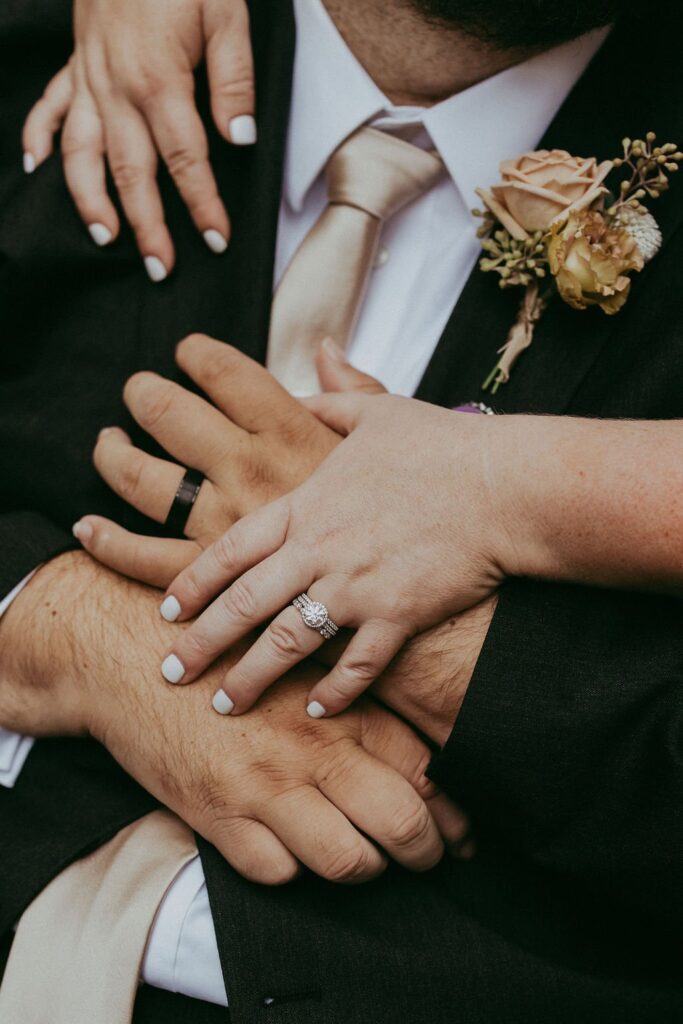 Close-up of a couple's hands with wedding rings, resting on the groom's chest. The groom wears a dark suit and beige tie, with a boutonniere featuring a peach rose.