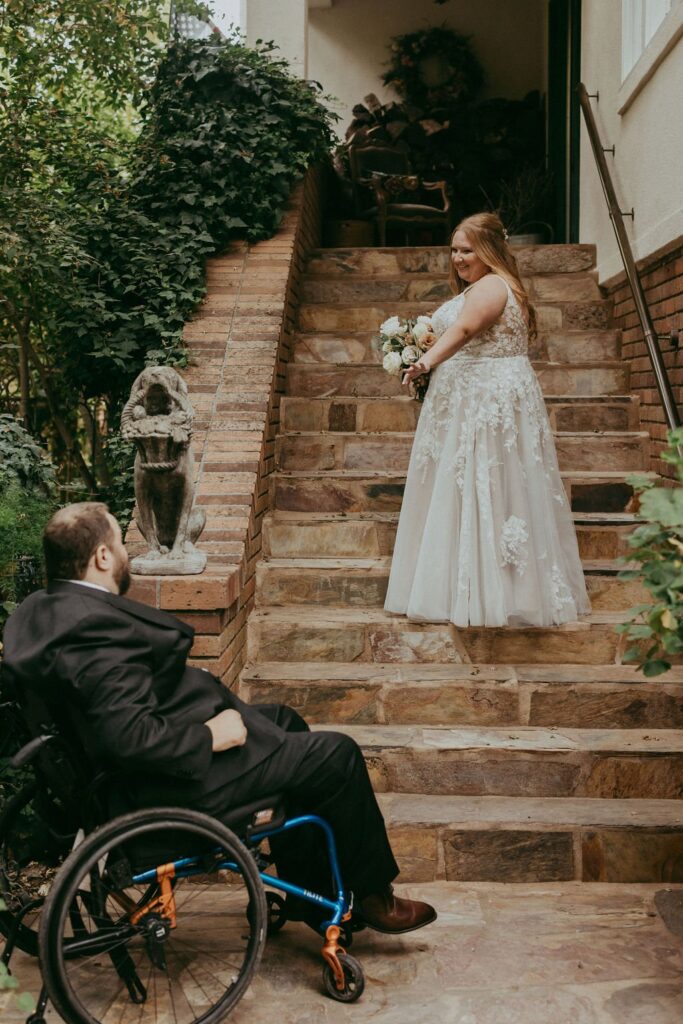 A woman in a wedding dress stands at the top of a stone staircase, looking back at a man in a wheelchair at the bottom, with greenery surrounding them.