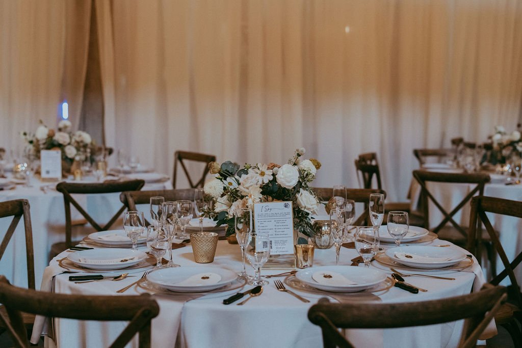 A wedding reception table with elegant place settings, floral centerpiece, candles, and a menu. In the background, similarly set tables are visible.