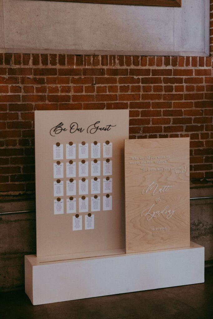 Two beige display boards stand against a brick wall. The left has organized cards labeled "Be Our Guest," and the right features a romantic quote with names and a date.