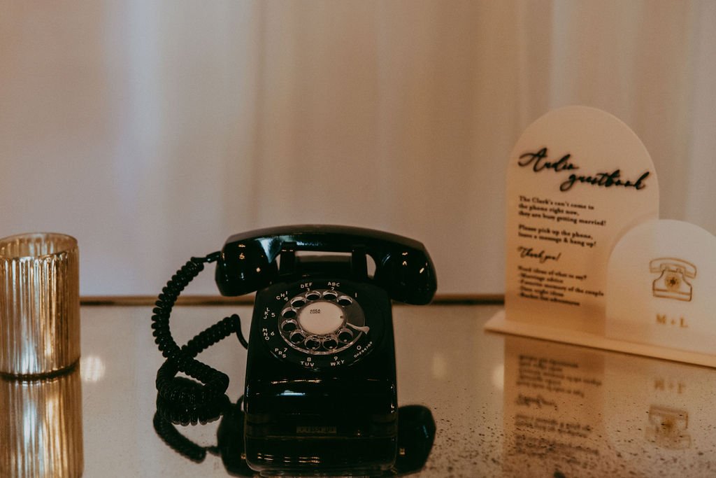 A black rotary phone on a glass surface next to a lit candle and a printed card with text.