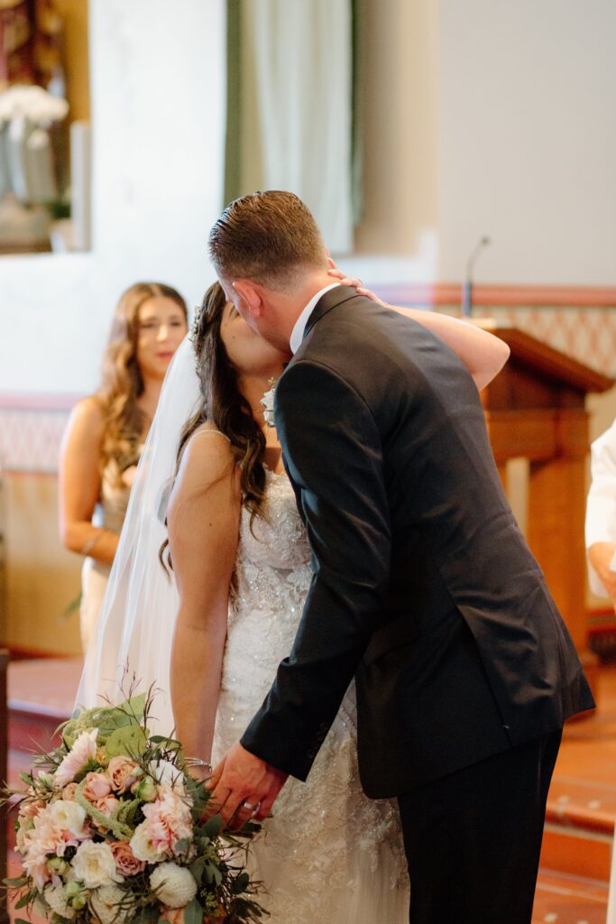 A bride and groom share a kiss at their wedding ceremony. The bride holds a bouquet of flowers. A bridesmaid stands in the background.