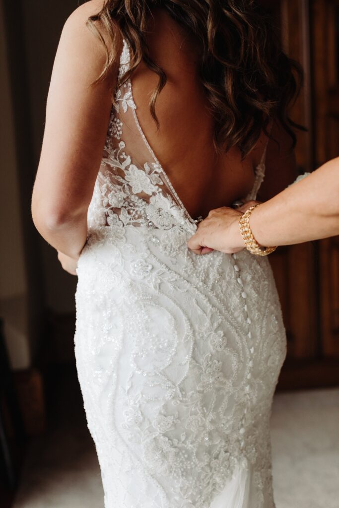 A woman wearing a lace wedding dress is being assisted by another person adjusting the dress from behind.