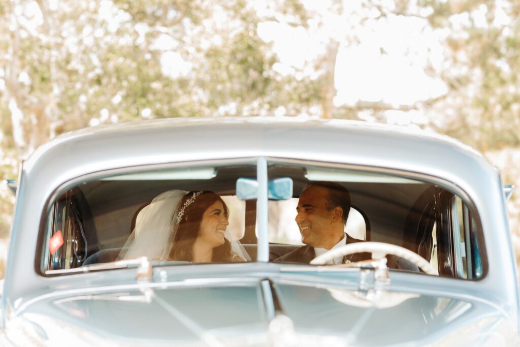A bride and a man smiling at each other inside a vintage car.