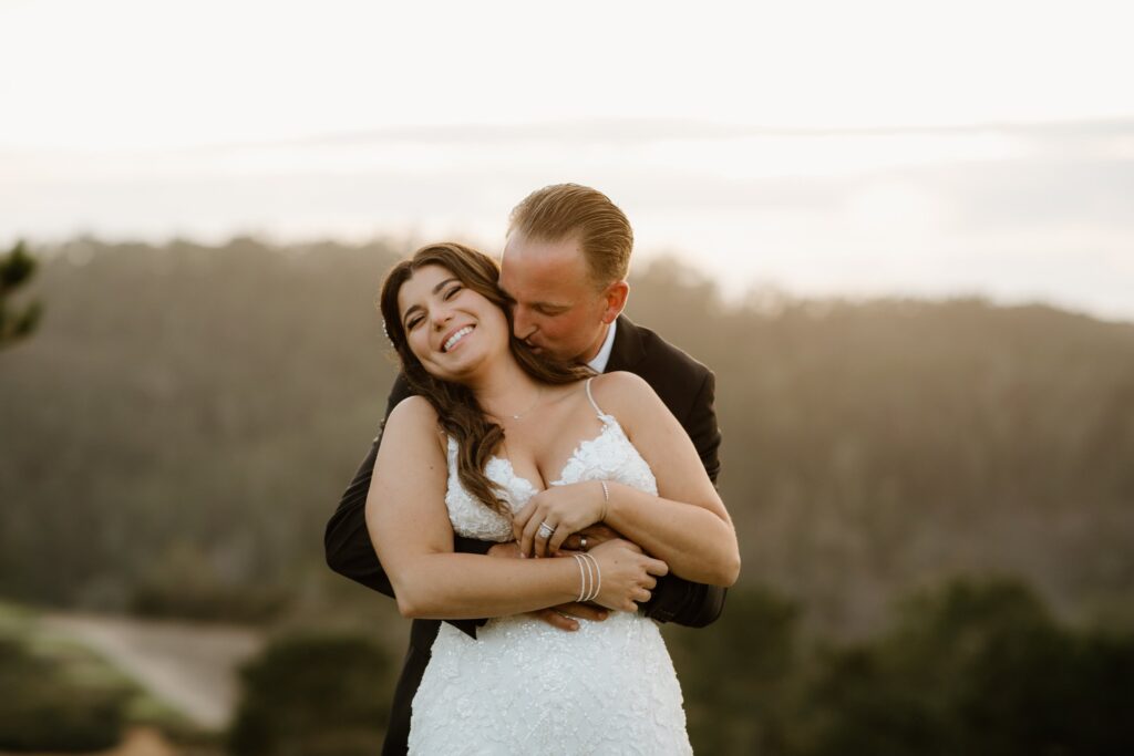 A couple embraces outdoors, with the woman in a white dress and the man in a dark suit, set against a blurred natural background.