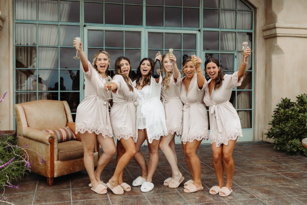 A group of six women in matching robes, one in white and the others in beige, holding drinks and smiling in a celebratory pose outdoors.
