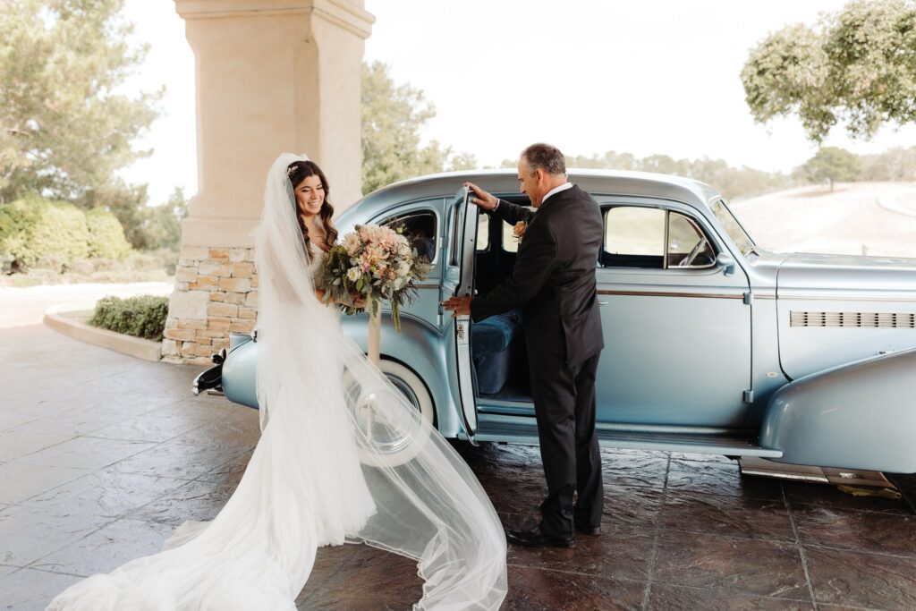 Bride in a white gown stands beside a classic blue car, holding a bouquet. A man in a suit opens the car door. Trees and stone pillars are in the background.