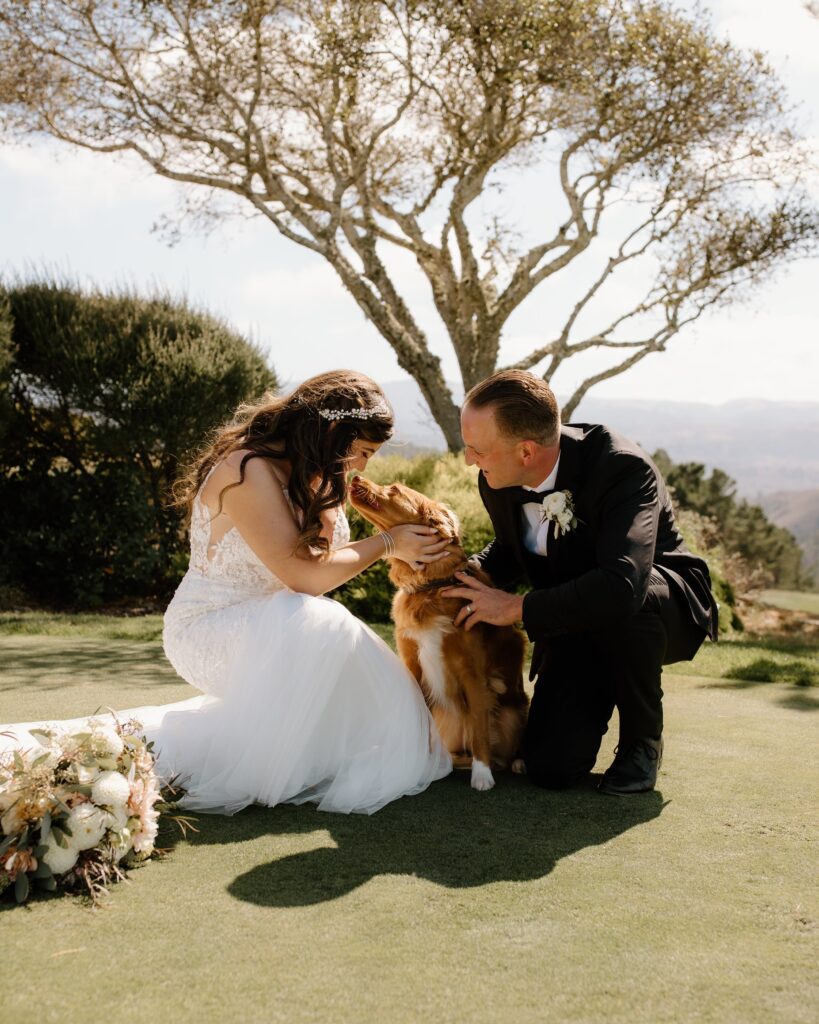 Bride and groom kneeling on grass petting a brown dog, with a bouquet nearby and a tree in the background.