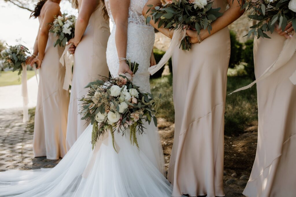 A bridal party stands outdoors holding bouquets, with the bride in a white gown and bridesmaids in beige dresses.