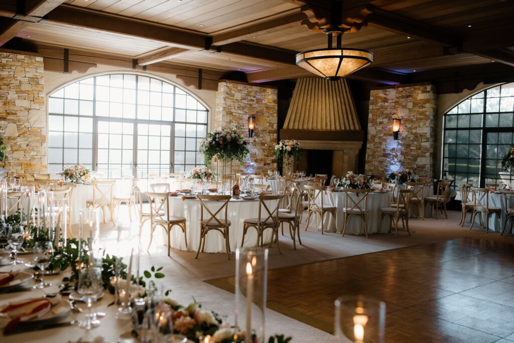 Elegant wedding reception hall with round tables, white tablecloths, floral centerpieces, and a large stone fireplace under a wooden ceiling.