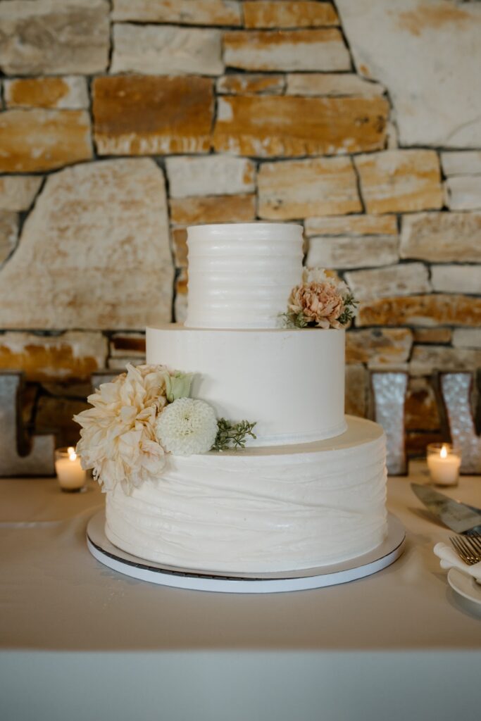 Three-tier white cake decorated with flowers, against a stone wall background.
