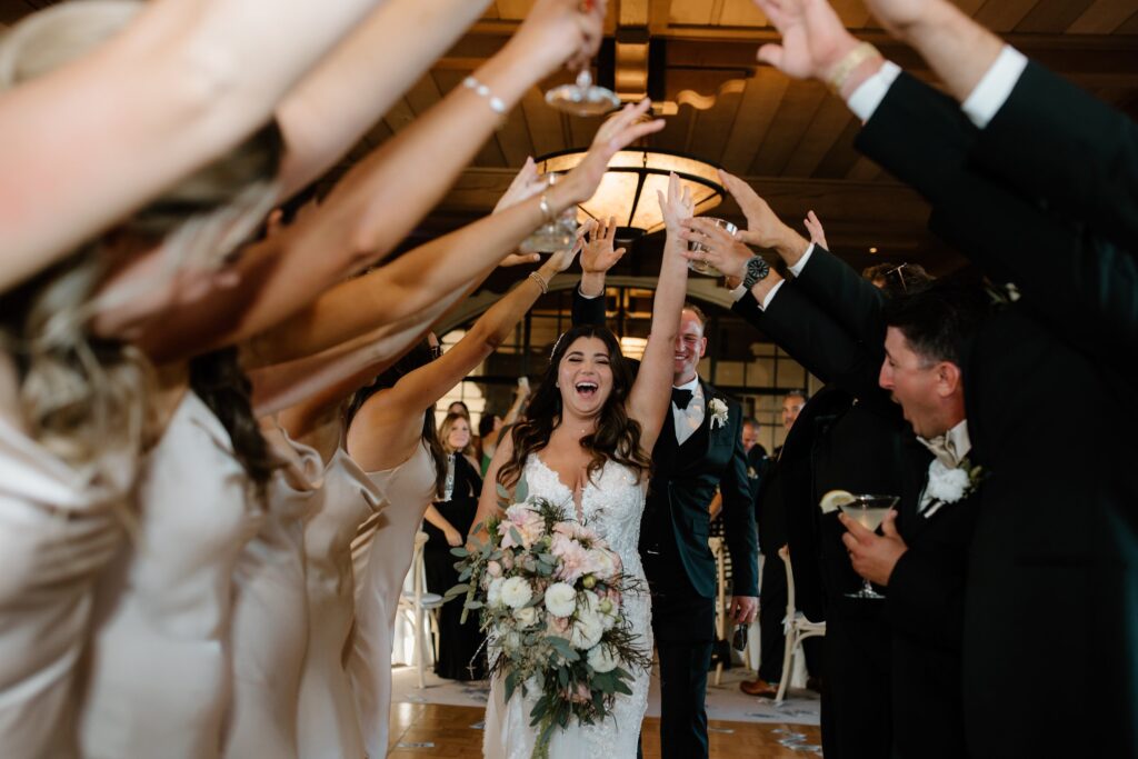 Bride and groom walk joyfully through a tunnel formed by guests' raised arms at a wedding reception. Bride holds a floral bouquet.