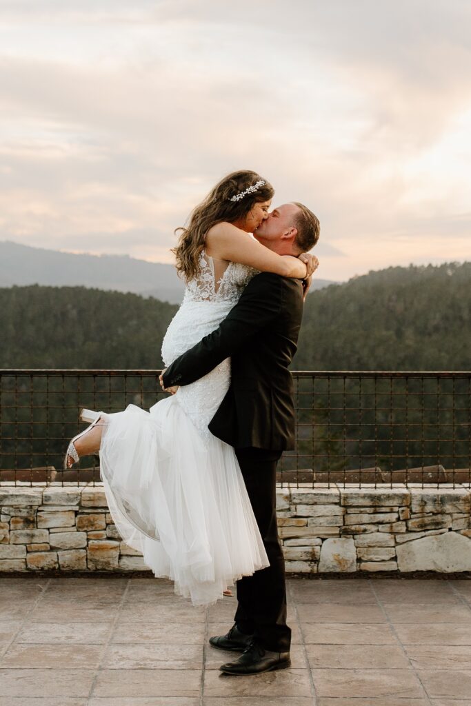 A groom lifts a bride in a white dress while they kiss, set against a scenic mountain backdrop with a stone railing.