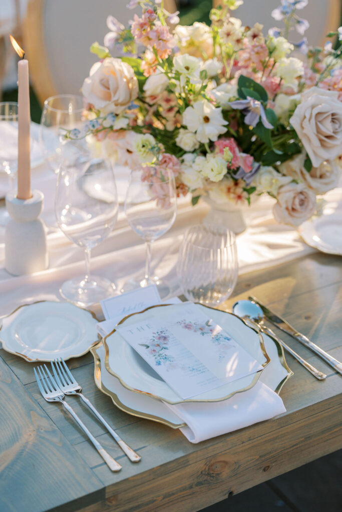Elegant table setting with floral centerpiece, gold-rimmed plates, silverware, and a lit candle on a wooden table.
