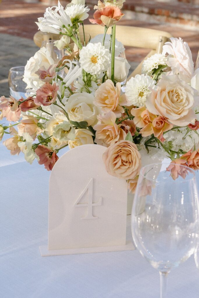 Elegant floral centerpiece with roses and assorted flowers on a table, accompanied by a table number card displaying the number "4," with empty wine glasses nearby.