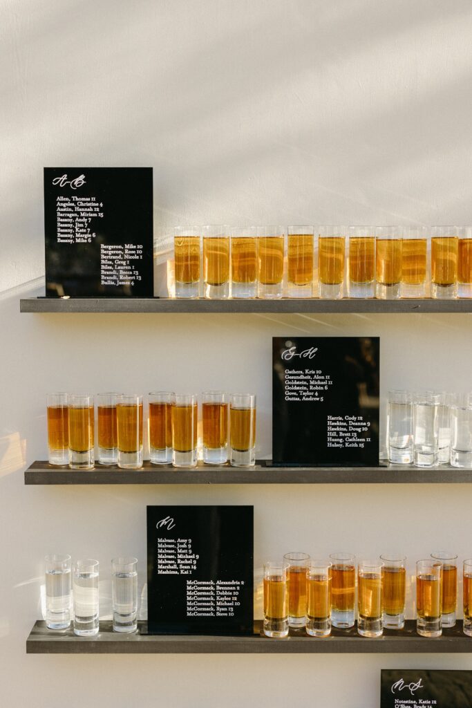 Three shelves display rows of filled shot glasses. Each shelf includes a black sign with a list of ingredient names.