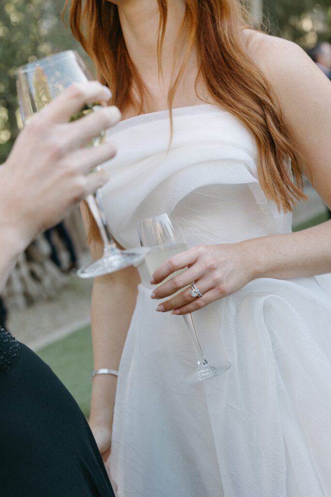 Two people holding champagne flutes, one wearing a white dress and the other in black attire, possibly at an outdoor gathering.