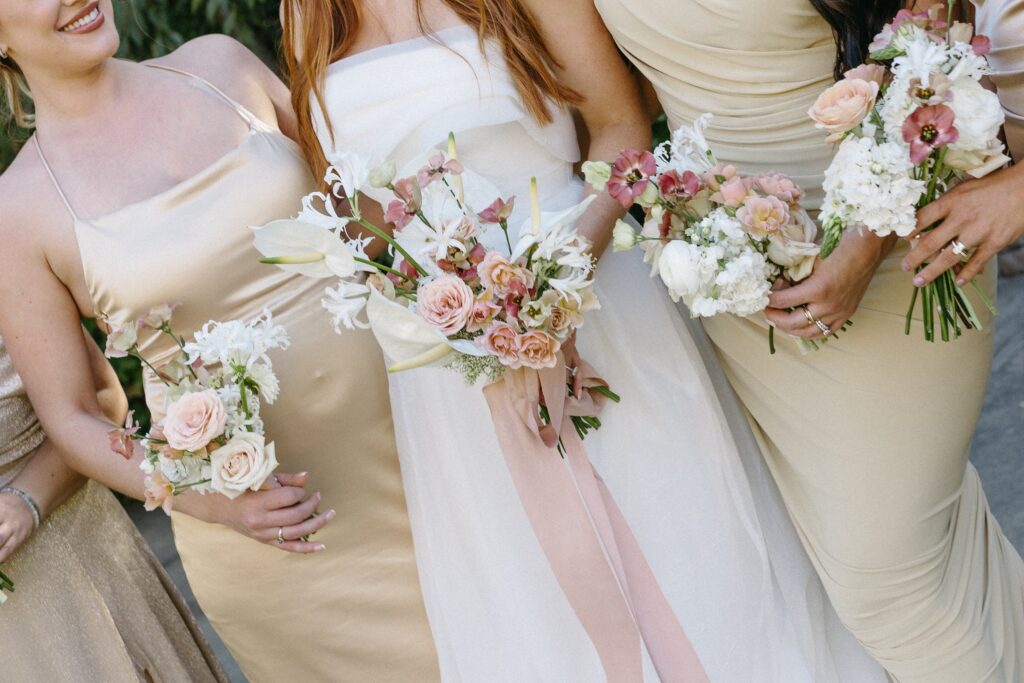 Three women in light-colored dresses hold bouquets of pastel flowers.