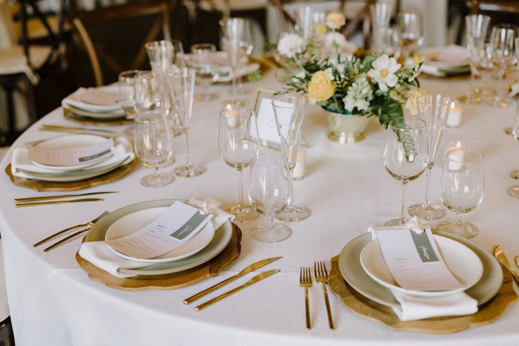 Elegant table setting with neatly arranged plates, gold cutlery, wine glasses, and a centerpiece with white and yellow flowers on a white tablecloth.
