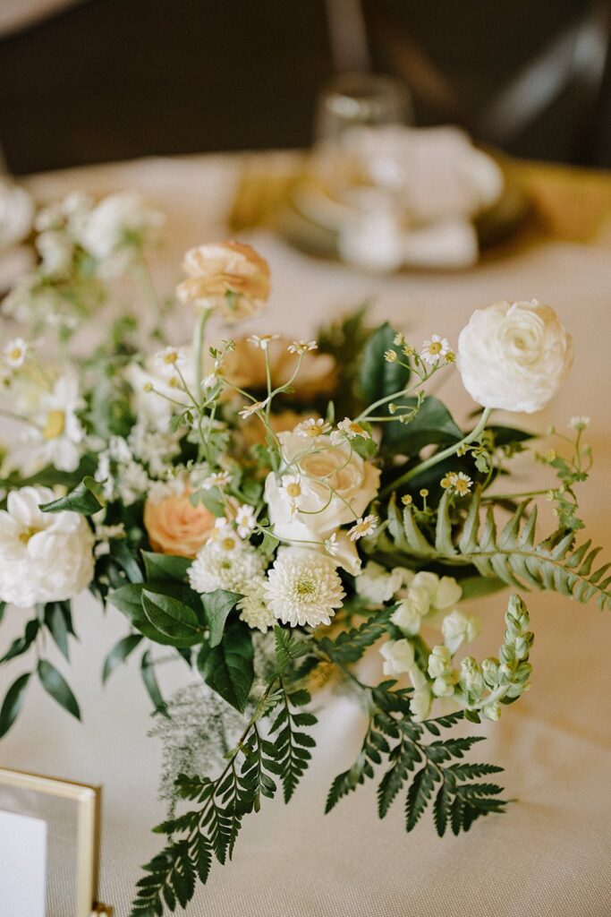 A floral arrangement with white and peach flowers, greenery, and ferns on a table.